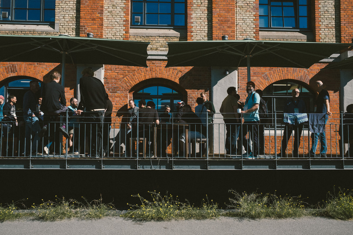 People standing on a sunny terrace with sunshades in front of a nice brick building.